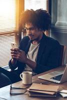 Important message. Young handsome African man using his smartphone while sitting at his working place photo