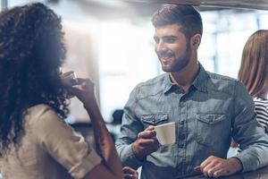 Coffee break with joy. Young handsome man holding coffee cup and discussing something with young woman while standing at bar counter photo
