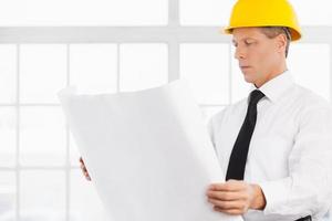 Architect at work. Confident senior man in formalwear and hardhat holding a blueprint and looking at it photo