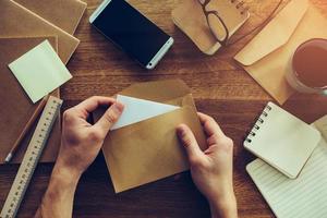 Opening envelope. Close-up top view of male hands opening envelope over wooden desk with different chancellery stuff laying on it photo