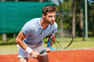 concentrado en el juego. un joven apuesto con camisa de polo sosteniendo una raqueta de tenis y luciendo concentrado mientras estaba de pie en la cancha de tenis foto