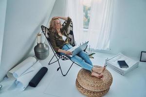 Attractive young woman using laptop while sitting in comfortable chair at home photo