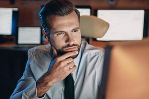 Thoughtful young man looking at the computer while staying late in the office photo