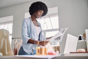 Beautiful young African woman analyzing fashion design sketch while standing near her desk in office photo