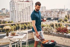 Handsome young man in casual clothing preparing barbecue while standing on the rooftop patio photo