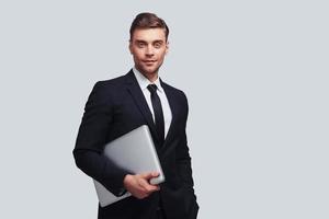 Pure confidence. Good looking young man in full suit carrying laptop and smiling while standing against grey background photo