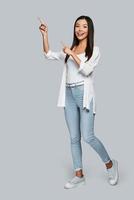 Look over here Full length of young Asian woman pointing copy space and smiling while standing against grey background photo