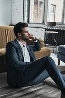 Enjoying quite minutes. Handsome young man in full suit and looking away while sitting on the floor at home photo