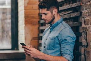 Message to her.Side view of handsome thoughtful young man holding smart phone and looking at it while leaning to brick wall in loft interior photo
