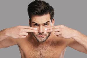 Taking good care of his skin. Handsome young man applying moisturizer and looking at camera while standing against grey background photo