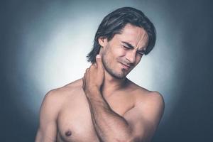 Feeling pain in neck. Portrait of young shirtless man keeping eyes closed and expressing negativity while touching his neck and standing against grey background photo