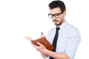 Making some urgent notes. Confident young man in shirt and tie making notes in his pad while standing against white background photo