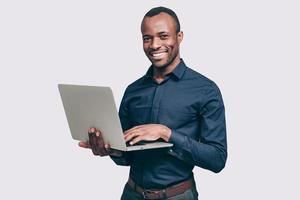 How may I help you Handsome young African man working on laptop and smiling to you while standing against grey background photo