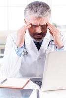 Stressed and tired doctor. Depressed mature grey hair doctor touching his head with hands while sitting at his working place photo