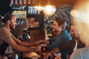 vista superior de jóvenes sonrientes con ropa informal bebiendo cerveza y uniéndose mientras están sentados en el pub foto