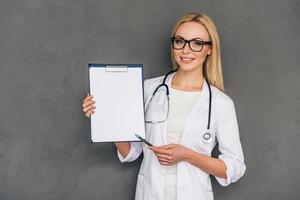 la mejor receta para usted hermosa joven doctora señalando su portapapeles y mirando la cámara con una sonrisa mientras está de pie contra un fondo gris foto