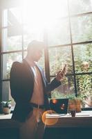 Typing quick business message. Low angle view of confident young man holding smart phone and looking at it while standing in front of the big window in office or cafe photo