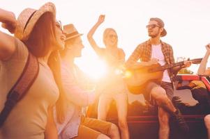 disfrutando de un viaje por carretera con los mejores amigos. grupo de jóvenes alegres bailando y tocando la guitarra mientras disfrutan juntos de su viaje por carretera en un camión de recogida foto