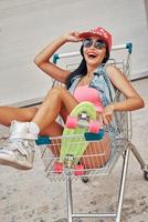 Living and loving this life Top view of happy young woman adjusting her cap and looking at camera while sitting in shopping cart against the garage door photo