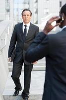 Business people. Rear view of African man in formalwear talking on the mobile phone while another businessman walking by stairs on background photo