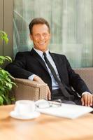 Taking a rest after business meeting. Cheerful mature man in formalwear looking at camera and smiling while sitting at the chair outdoors with coffee cup on foreground photo