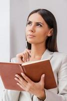 Waiting for inspiration. Thoughtful young businesswoman in suit holding note oad and touching her chin with pen while leaning at the wall photo