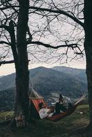 Better than any spa. Handsome young man lying in hammock and reading a book while camping with his girlfriend photo