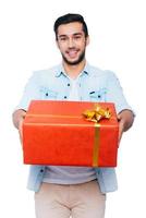 This is for you Happy young Indian man stretching out red gift box and smiling while standing against white background photo