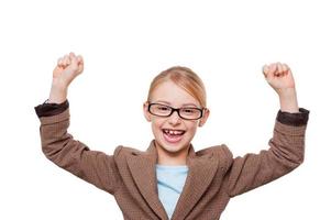 Yes Cheerful little girl in formalwear keeping her arms raised and smiling while standing isolated on white photo