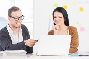 Working on the project together. Beautiful woman holding hand on chin and looking at the laptop while cheerful young man pointing on screen and smiling photo