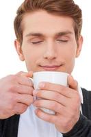 Drinking fresh coffee. Handsome young man holding cup of coffee and keeping eyes closed while standing isolated on white photo