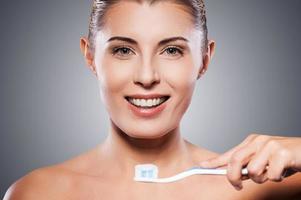 cuidado de los dientes hermosa mujer madura sosteniendo un cepillo de dientes cerca de su boca y sonriendo a la cámara mientras se enfrenta a un fondo gris foto