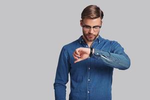 Time is money.  Thoughtful young man in eyewear checking the time while standing against grey background photo