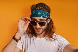 Self portrait of charming young man looking at camera and adjusting eyewear while standing against yellow background photo