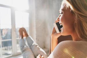 Rear view of beautiful young woman talking on her smart phone and smiling while sitting near the window at home photo