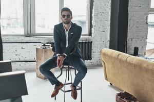 He got style. Handsome young man in full suit and eyewear keeping hands clasped while sitting on the stool photo