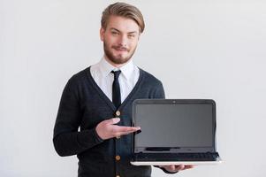 su publicidad aquí apuesto joven sosteniendo una computadora portátil y apuntándola mientras está de pie contra un fondo gris foto