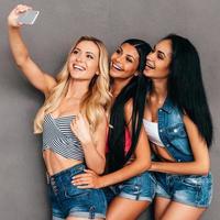 Friendly selfie. Side view of three beautiful women bonding to each other and smiling while making selfie against grey background photo