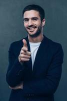 You are awesome Cheerful young man pointing you and looking at camera while standing against grey background photo