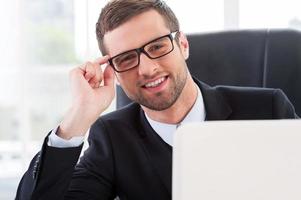 siempre dispuesto a ayudarte. un joven alegre con ropa formal mirando por la laptop y ajustando sus lentes mientras se sienta en su lugar de trabajo foto