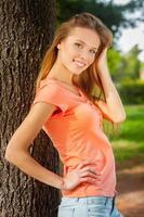 Beauty in nature. Side view of beautiful young woman holding hand in hair and smiling while leaning at the tree photo