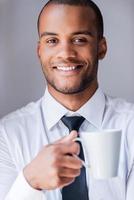 Relaxing during coffee break. Handsome young African man in shirt and tie holding cup with hot drink and smiling photo