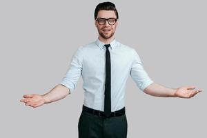 Handsome young man in formalwear looking at camera and keeping arms outstretched while standing against grey background photo