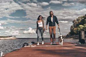 Carefree day together. Full length of beautiful young couple walking with their dog on the river shore while spending time outdoors photo