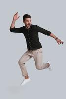 Feeling free to do anything. Full length of playful young man gesturing and smiling while jumping against grey background photo