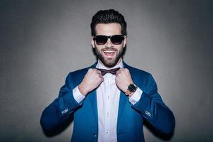 Always in good mood Close-up of handsome young cheerful man wearing sunglasses adjusting his bow tie and looking at camera with smile while standing against grey background photo