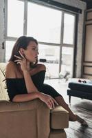 Charming beauty. Attractive young woman in elegant black dress keeping hand in hair and looking away while sitting on the sofa photo