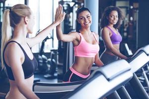 We did it Side view of young beautiful women looking at each other and giving high-five with smile while running on treadmill at gym photo