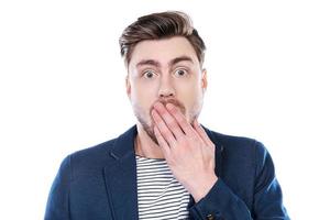 It is unbelievable Portrait of surprised young man looking at camera and covering mouth with hand while standing against white background photo