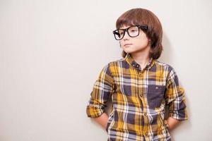 lleno de inspiración. niño pequeño con anteojos apoyado en la pared y mirando hacia otro lado mientras está de pie contra un fondo gris foto